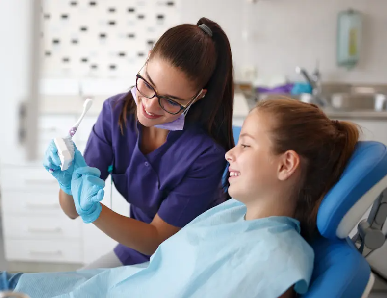 Dental assistant showing a young patient how to brush her teeth in a dentist office 