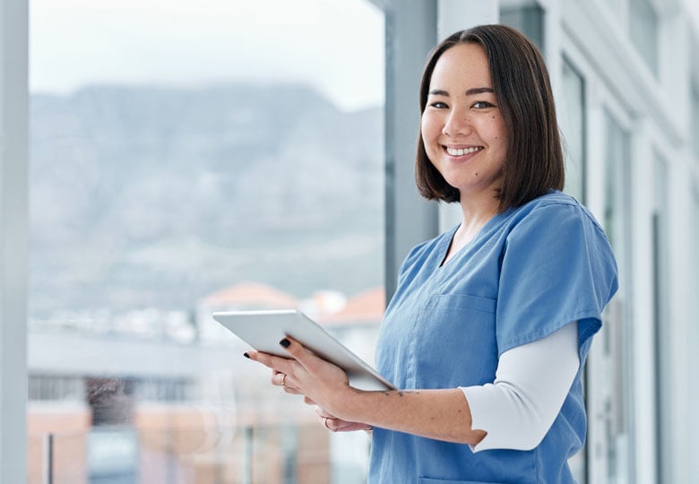 A registered nurse standing by a hospital window holding a tablet and smiling