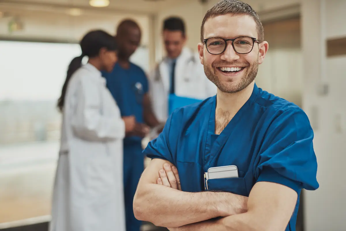 A male nurse smiling with his arms crossed with several other nurses and physicians standing in the background