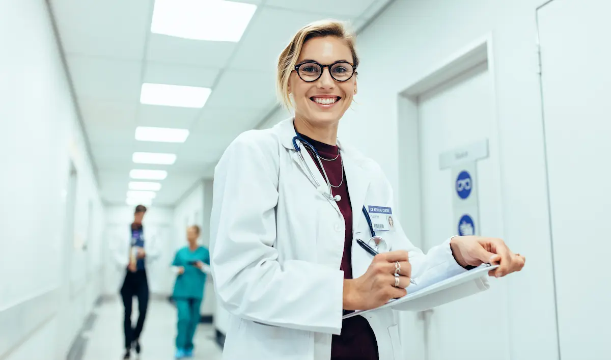 A nurse practitioner holding a clipboard standing in the hallway of a hospital