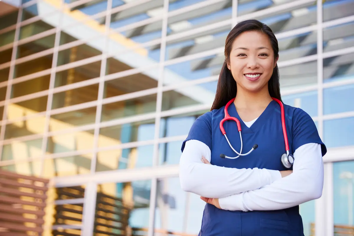 A registered nurse standing in front of a healthcare facility smiling with her arms crossed
