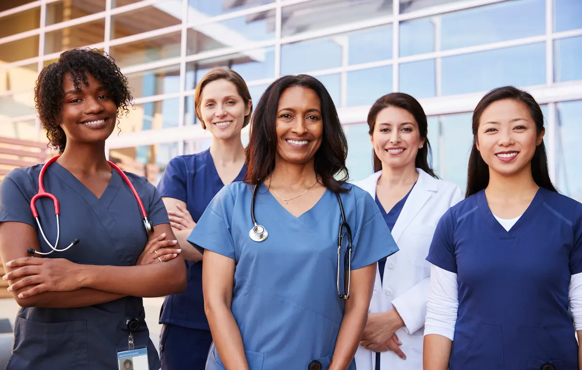 Four female registered nurses and a female physician smiling while standing in front of a healthcare facility