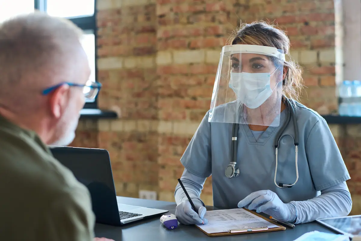 Front line worker conducting a COVID screening wearing a face shield at a desk with an older male patient