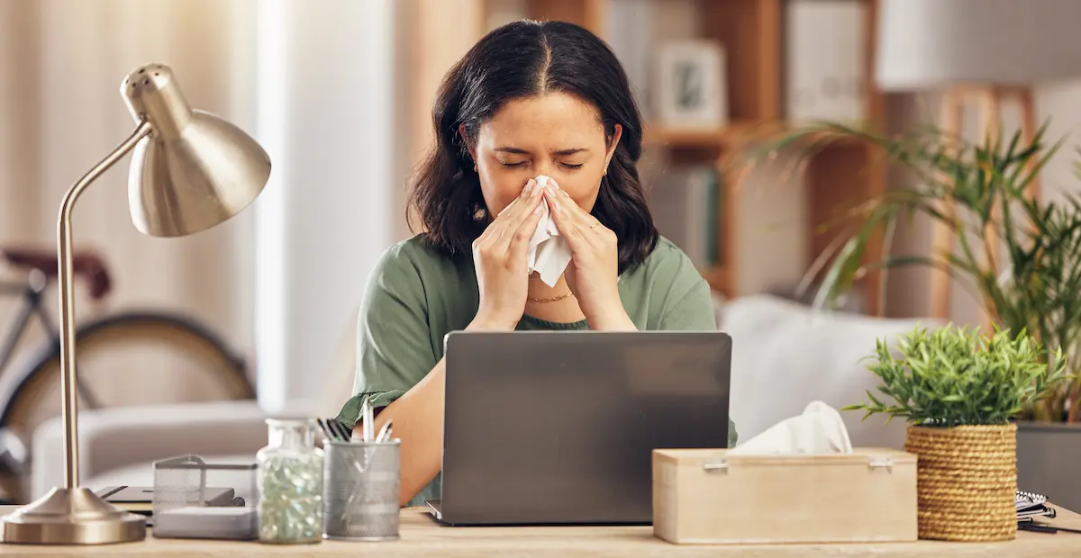 Woman sick with the flu sneezing into a tissue while working at home