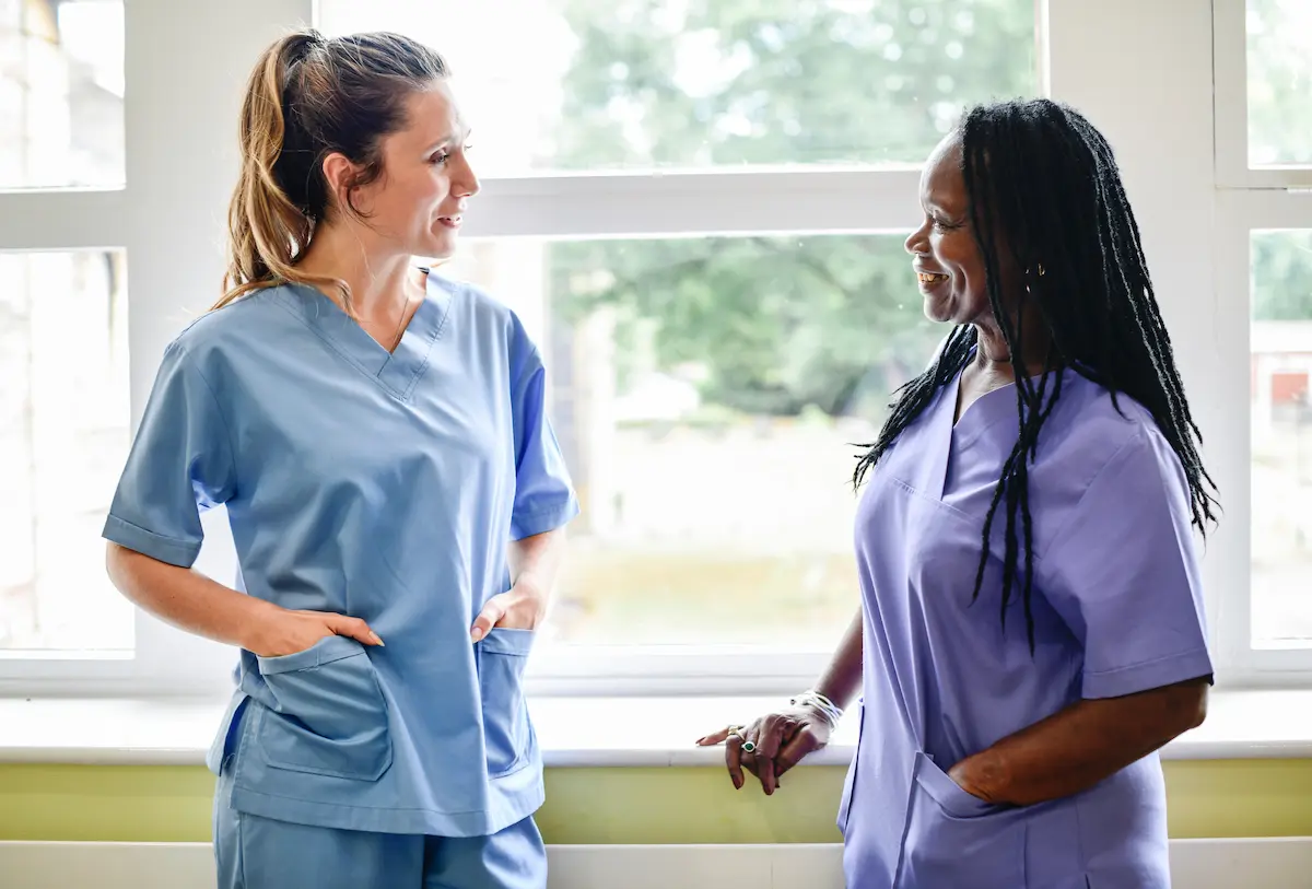 Two female registered nurses standing in the hallway of a hospital smiling at each other while having a conversation